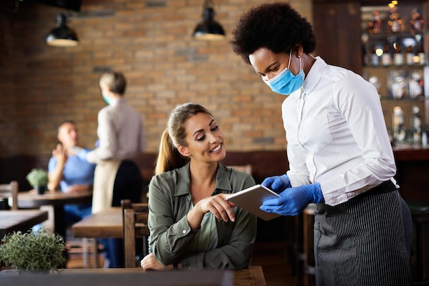 Garçonete negra com máscara facial fazendo pedido no touchpad de um cliente em um café