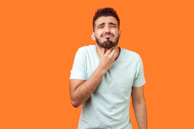 Garganta inflamada Retrato de homem moreno doente com barba em camiseta branca tocando pescoço doloroso sofrendo dor de garganta sintoma de amigdalite conceito médico estúdio tiro isolado em fundo laranja