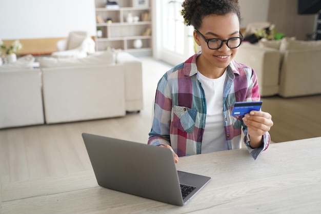 Garota adolescente afro-americana mantém compras de cartão de crédito bancário on-line no laptop em casa e-commerce