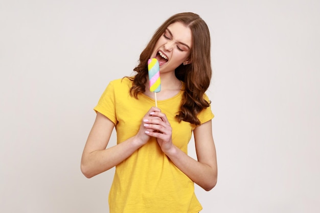 Garota adolescente animada na camiseta amarela estilo casual mordendo sorvete tem expressão louca desejando comer com grande prazer deseja fome Tiro de estúdio interno isolado em fundo cinza