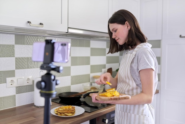 Garota adolescente, blogueira de comida cozinhando panquecas em casa na cozinha, filmando receita de vídeo. Fêmea com laranja perto do fogão. Hobbies, canal de vídeos com seguidores, crianças e adolescentes