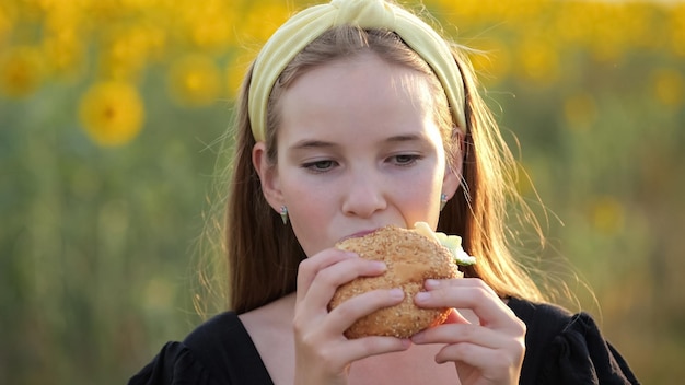 Garota adolescente come hambúrguer contra campo de girassol embaçado