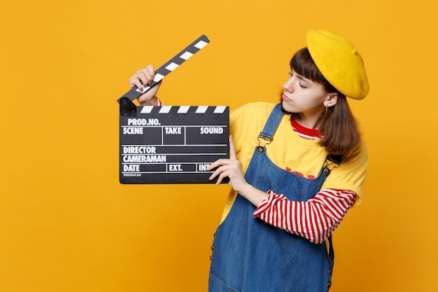 Garota adolescente de boina francesa e vestido de denim olhando no clássico filme preto fazendo claquete isolada no fundo da parede amarela. Conceito de estilo de vida de emoções sinceras de pessoas. Mock up espaço de cópia.