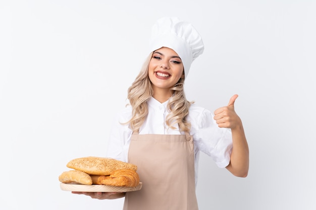 Garota adolescente em uniforme de chef. Padeiro feminino segurando uma mesa com vários pães sobre branco isolado, dando um polegar para cima gesto