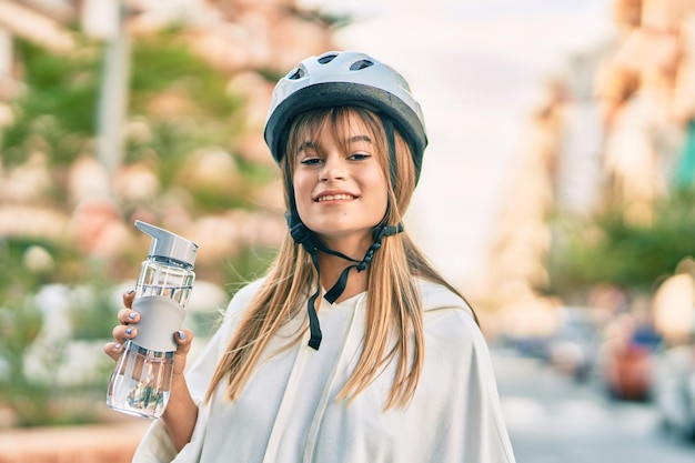 Garota adolescente esportiva caucasiana usando capacete de bicicleta e bebendo garrafa de água na cidade
