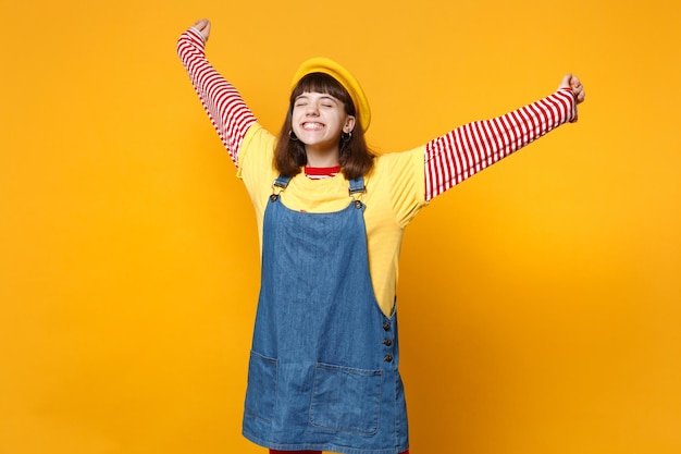 Garota adolescente feliz na boina francesa, vestido jeans, mantendo os olhos fechados, espalhando e levantando as mãos isoladas no fundo da parede amarela. Emoções sinceras de pessoas, conceito de estilo de vida. Mock up espaço de cópia.