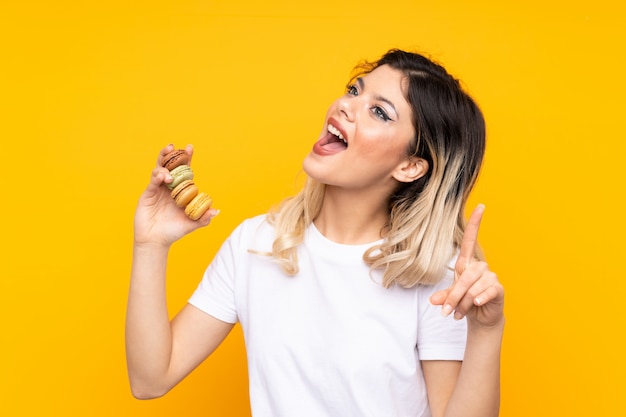 Garota adolescente isolada na parede amarela segurando macarons franceses coloridos e apontando uma ótima idéia