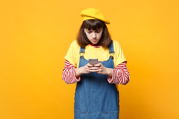 Garota adolescente preocupada em boina francesa, vestido jeans usando telefone celular, digitando mensagem sms isolada em fundo amarelo no estúdio. Emoções sinceras de pessoas, conceito de estilo de vida. Mock up espaço de cópia.