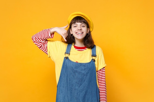 Garota adolescente rindo em vestido de denim de boina francesa fazendo gesto de telefone como diz me ligue de volta isolado em fundo amarelo no estúdio. Conceito de estilo de vida de emoções sinceras de pessoas. Mock up espaço de cópia.