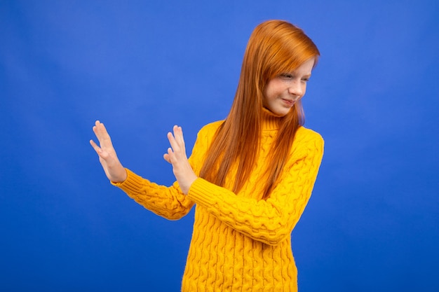 Garota adolescente ruiva europeia ofendida em um suéter amarelo acenou com as mãos em negação em azul