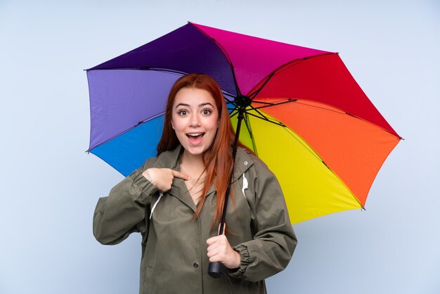 Garota adolescente ruiva segurando um guarda-chuva sobre parede azul isolada com expressão facial de surpresa