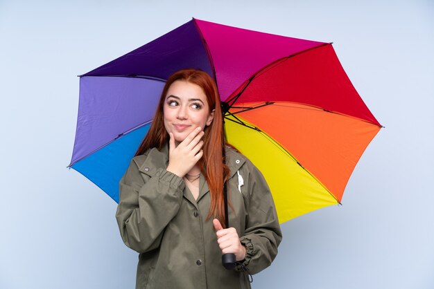 Garota adolescente ruiva segurando um guarda-chuva sobre parede azul isolada, pensando uma idéia