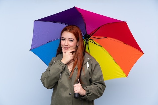 Garota adolescente ruiva segurando um guarda-chuva sobre parede azul isolada rindo