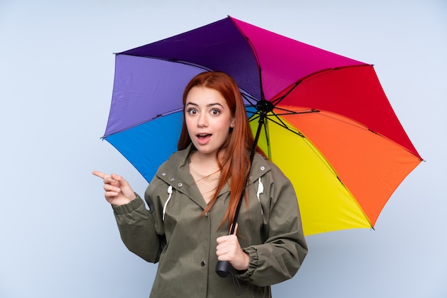 Garota adolescente ruiva segurando um guarda-chuva sobre parede azul isolada surpreendeu e apontando o dedo para o lado