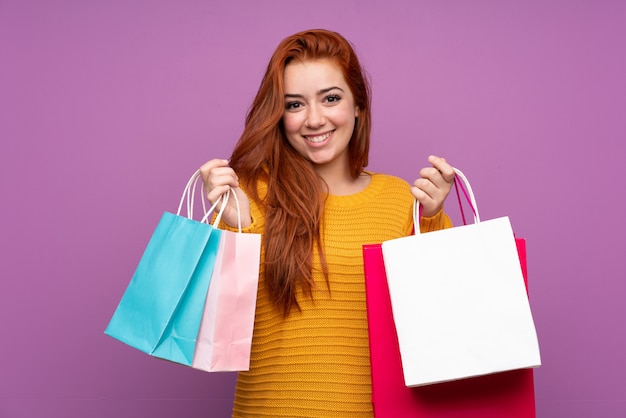 Garota adolescente ruiva sobre parede roxa isolada segurando sacolas de compras e sorrindo