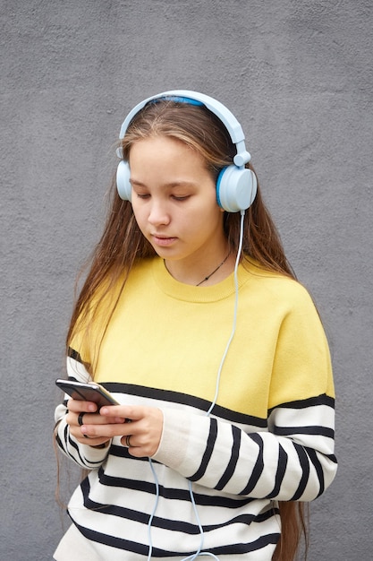 Foto garota adolescente séria em um suéter listrado amarelo usa um smartphone e fones de ouvido ouve música em um fundo cinza