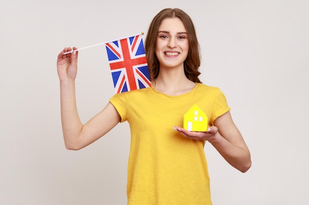 Garota adolescente sorridente autoconfiante em roupas de estilo casual, segurando a bandeira da Grã-Bretanha e a casa de papel, sonhando em comprar acomodação na Inglaterra. Tiro de estúdio interior isolado em fundo cinza.