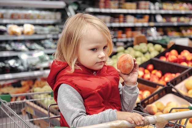 Garota adorável criança sentada no carrinho de compras em uma loja de frutas de alimentos ou supermercado. Retrato de criança fofa indo às compras e comprando frutas e vegetais frescos. Conceito de comida saudável para crianças