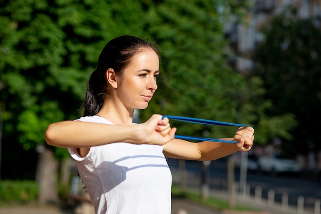 Garota adorável e esportiva fazendo exercícios de mãos com elástico azul no parque