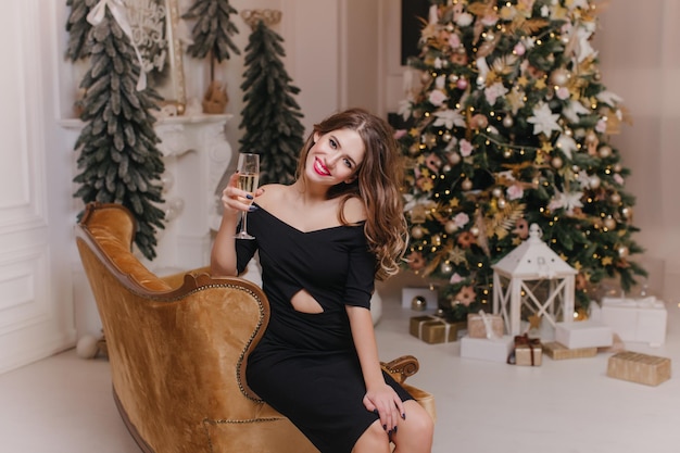 Garota adorável em um vestido preto elegante, levantando a taça de champanhe na frente da árvore de Natal. Adorável modelo feminino em um traje elegante, aproveitando a celebração do ano novo em casa.