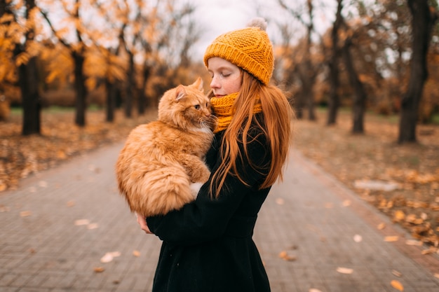 Garota adorável sardas com expressão de rosto gentil, segurando seu gato vermelho em um vale do parque outono.