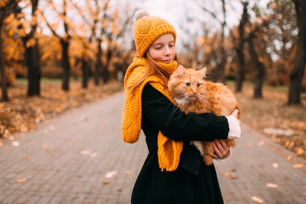 Garota adorável sardas com expressão de rosto gentil, segurando seu gato vermelho em um vale do parque outono.