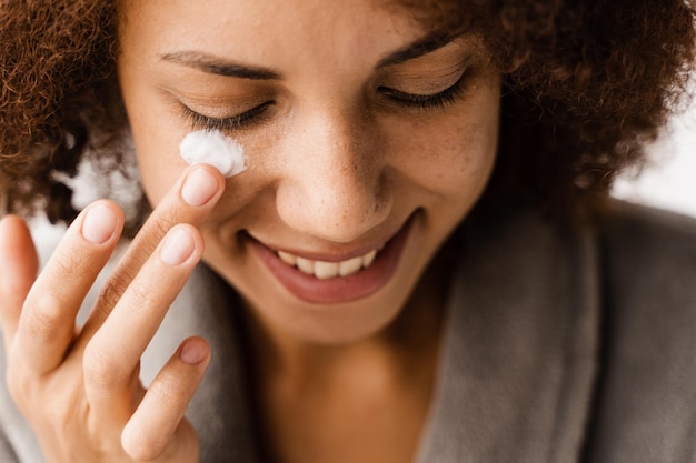 Foto garota africana aplicando creme hidratante facial closeup para proteger a pele da secura no banho mulher afro-americana em roupão de banho com creme hidratante facial fazendo rotina de beleza matinal