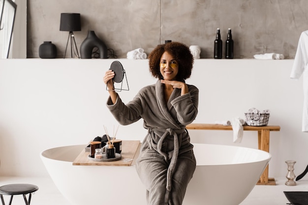 Garota africana em roupão de banho com tapa-olhos dourado está olhando para o espelho no banheiro Rotina de beleza matinal de mulher afro-americana