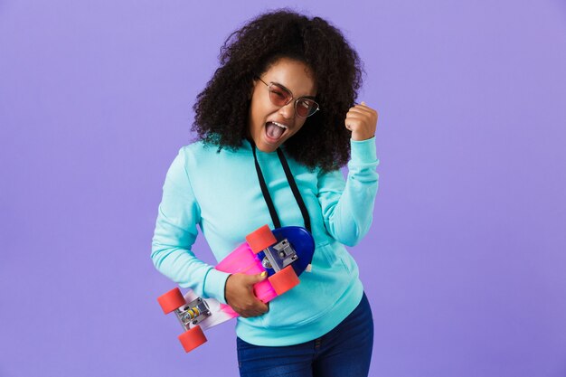 Garota africana posando isolado sobre o espaço violeta, segurando o skate, mostrando o gesto do vencedor.