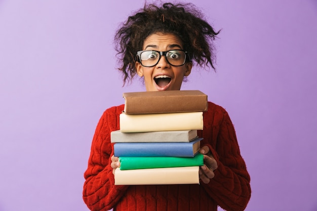 Garota afro-americana alegre em uniforme escolar segurando um monte de livros, isolado