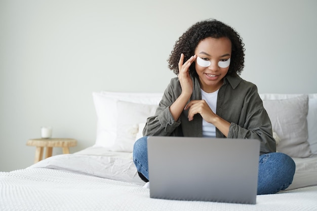 Garota afro-americana aplicando sob os tapa-olhos sentado no laptop na cama Rotina de beleza Skincare