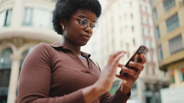 Garota afro-americana atraente usando óculos em pé na rua com smartphone Mulher de pele escura parecendo concentrada usando o telefone