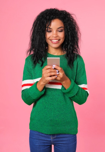 Garota afro-americana com um suéter verde com listras brancas e vermelhas está olhando para a tela de seu smartphone, que está segurando nas mãos, enquanto sorri sinceramente