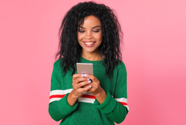 Garota afro-americana com um suéter verde com listras brancas e vermelhas está olhando para a tela de seu smartphone, que está segurando nas mãos, enquanto sorri sinceramente