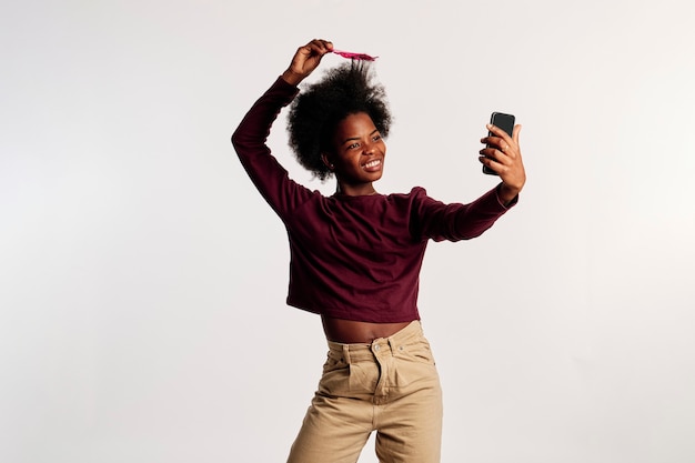 Garota afro-americana em poses de suéter marrom enquanto olha para o telefone e penteia o cabelo com uma escova rosa.