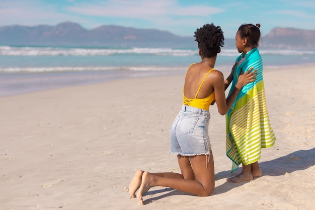 Garota afro-americana enrolada em toalha com jovem mãe olhando para o mar contra o céu em dia ensolarado