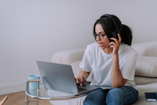 Garota afro-americana focada em camiseta branca e óculos sentado no sofá trabalha usando laptop em casa