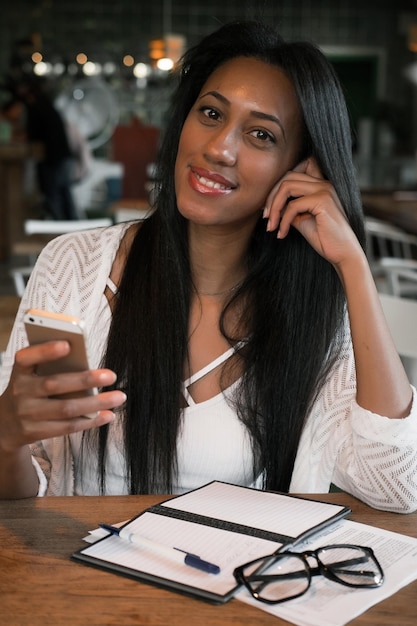 Garota afro-americana hipster escrevendo para fazer a lista e recebendo mensagem no smartphone Estudante sorridente fazendo lição de casa enquanto lê a notificação com boas notícias no celular
