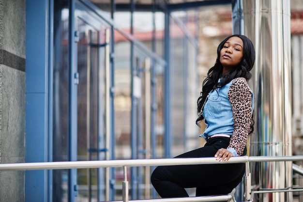 Garota afro-americana hipster vestindo camisa jeans com mangas de leopardo posando na rua contra prédio de escritórios moderno com janelas azuis