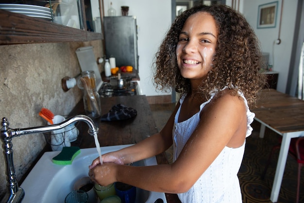 Garota afro-americana na cozinha de casa lavando a louça