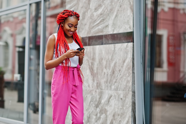 Garota afro-americana na moda em calças cor de rosa e temores vermelhos posou ao ar livre com telefone móvel.