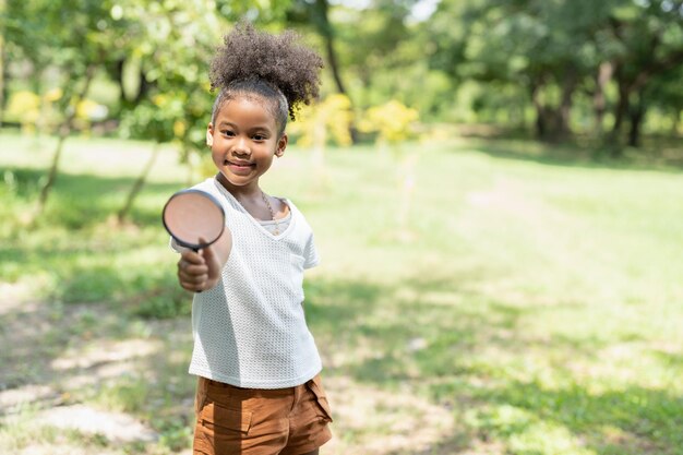 Garota afro-americana se diverte segurando lupa para explorar e ver insetos na árvore no parque