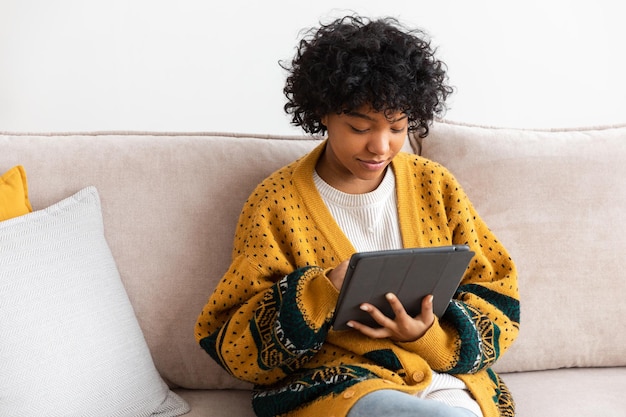 Garota afro-americana segurando a tela de toque do tablet digital digitando página de rolagem em casa Mulher com dispositivo de tecnologia móvel navegando na Internet usando aplicativos de mídia social jogando jogo Estudar trabalho fazer compras online