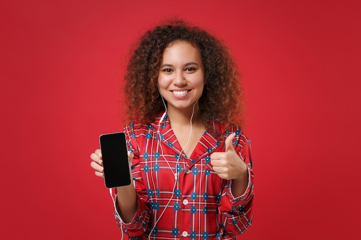 Foto garota afro-americana sorridente em homewear de pijama descansa em casa isolada em fundo vermelho. relaxe o estilo de vida de bom humor. ouça música com fones de ouvido segure o celular com a tela vazia mostrando o polegar para cima.