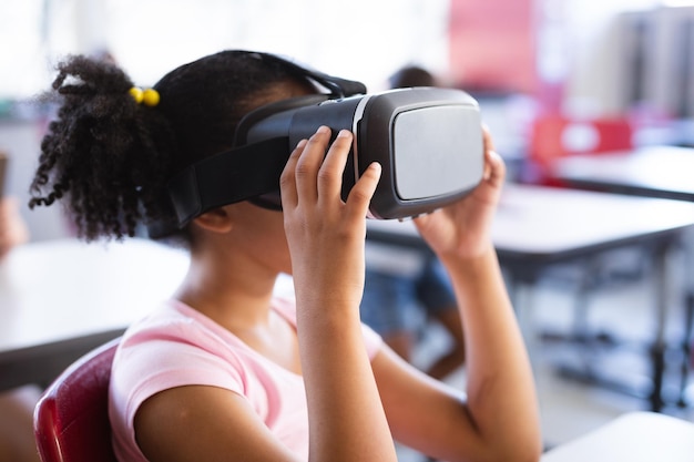 Garota afro-americana usando fone de ouvido vr enquanto está sentada em sua mesa na classe na escola