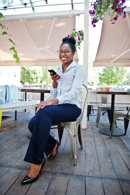 Garota afro-americana usar óculos com telefone celular sentado no caffe ao ar livre.