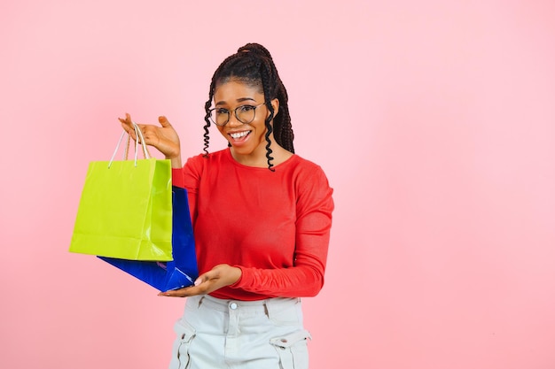 Garota afro animada de grande venda com sacolas de compras tocando óculos de sol olhando para a câmera sobre a parede do estúdio rosa