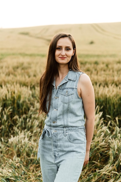 Garota agricultora. uma linda garota de macacão jeans fica entre um campo de trigo. espigas de trigo maduras. garota ao pôr do sol no campo