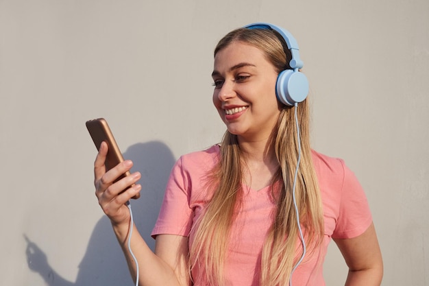 Garota alegre de camiseta rosa está falando em uma chamada de vídeo em fones de ouvido azuis olhando para a tela de um telefone celular