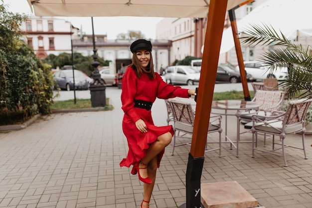 Garota alegre e brincalhona se move na rua Garota de cabelos castanhos em boné estiloso e vestido vermelho com olhares de sorriso